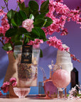 A serene still-life arrangement featuring The High Priestess Box, including a vase with pink blossoms and green leaves, a clear bag of High Priestess Wisdom Tea, a glass teapot, a pink glass orb, various crystals, and a small bowl, all set against a gradient blue-to-purple background.