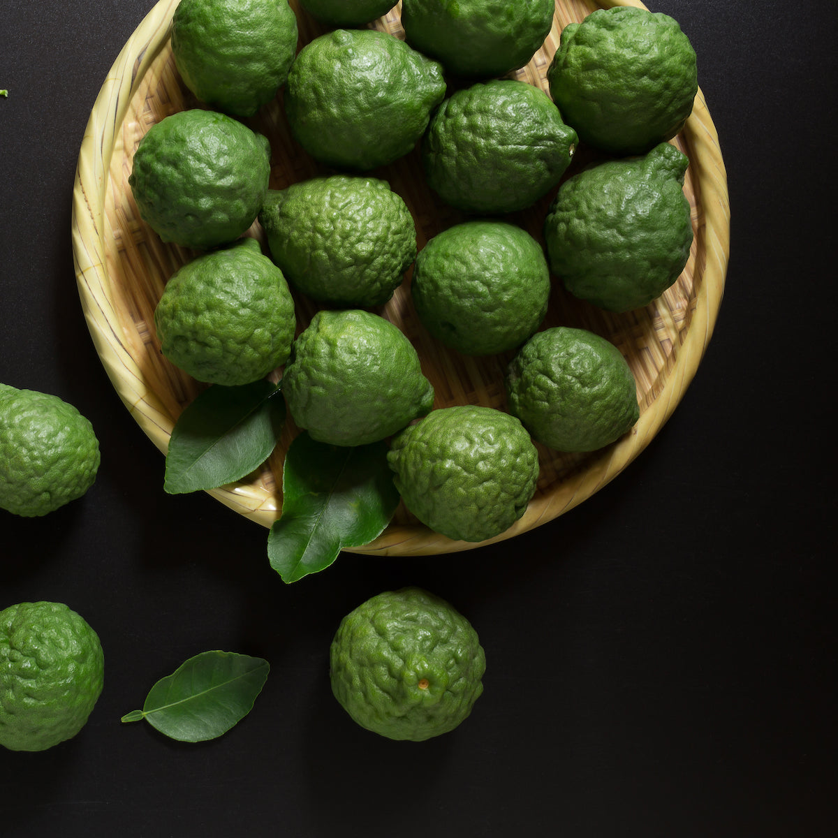 A woven bamboo plate is filled with green kaffir limes. Some limes and a few leaves are scattered on a black surface around the plate. The limes have a bumpy texture and vibrant green color.