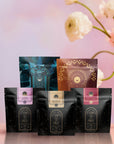 Five Bestsellers of Magic Tea Starter Set packages, with black and gold designs up front and colorful wellness blends behind, are arranged on a pastel floral background. White flowers blur in the distance, highlighting this 60-75 cup sampler box.