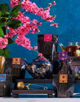 A colorful display features various bags of Magic Hour's Black Tea Magic Sampler Box - Bestselling Organic Black Teas for Energy & Vitality on a dark wooden rack, with a dark blue teapot adorned with golden patterns in the center. A gold tea infuser and a glass teapot with organic tea are nearby. Pink cherry blossoms in a pot enhance the vibrant setting.
