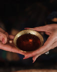 Close-up of a person's hands holding a small, ornately decorated brass cup filled with tea. The person's fingers have polished nails, and they are wearing a beaded bracelet and a ring with a large stone. The background is softly blurred and dark.