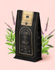 A package of Bliss: Sacred Tulsi Adaptogen Tea, adorned with gold accents and the “Magic Hour” label, stands before illustrated green leaves and purple flowers on a pale pink background, evoking the calming benefits of Tulsi (Holy Basil), celebrated for its natural sleep aid qualities.