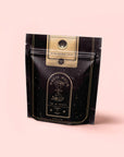 A black and gold resealable pouch labeled “Bliss: Sacred Tulsi Adaptogen Tea” with an hourglass design, set against a light pink background. The package features celestial and infinity-themed graphics, highlighting Tulsi (Holy Basil) as a natural sleep aid.