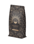 The "Breakfast with Benefits: Blood Sugar Balance" coffee bag boasts golden lotus flowers and star patterns on a dark background. Infused with Assam tea notes, it aims to make mornings magical while supporting blood sugar balance.