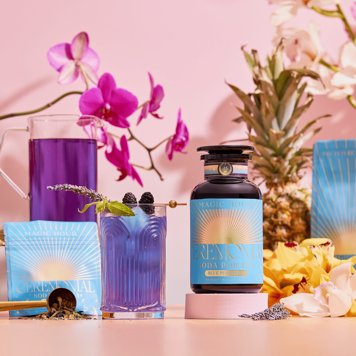 A blue-purple drink made with blackberries, herbs, and Butterfly Pea Flower stands next to a Blue Pineapple: Ceremonial Soda Pop Tea bottle and a sachet. A glass pitcher, flowers, and tea scoop enhance the pastel pink scene.