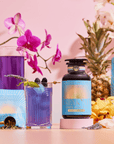 A blue-purple drink made with blackberries, herbs, and Butterfly Pea Flower stands next to a Blue Pineapple: Ceremonial Soda Pop Tea bottle and a sachet. A glass pitcher, flowers, and tea scoop enhance the pastel pink scene.