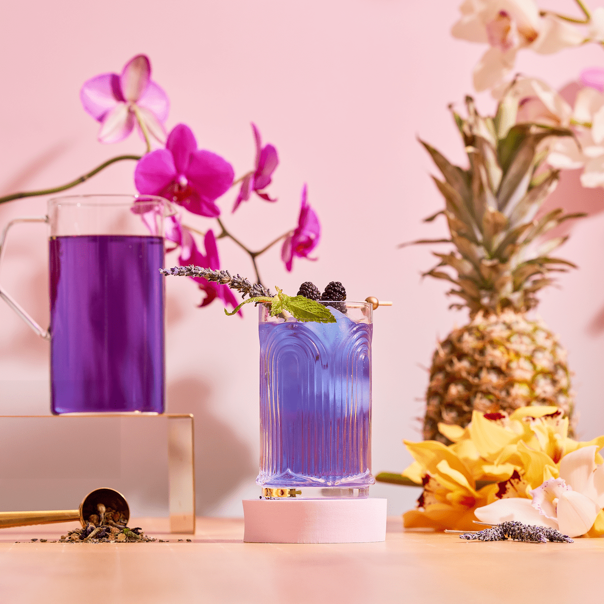 A glass of Blue Pineapple: Ceremonial Soda Pop Tea, made with antioxidant-rich tea and garnished with lavender, blackberries, and mint, sits beside a matching pitcher amid orchids, pineapple, yellow flowers, and a tea scoop on a pink background.