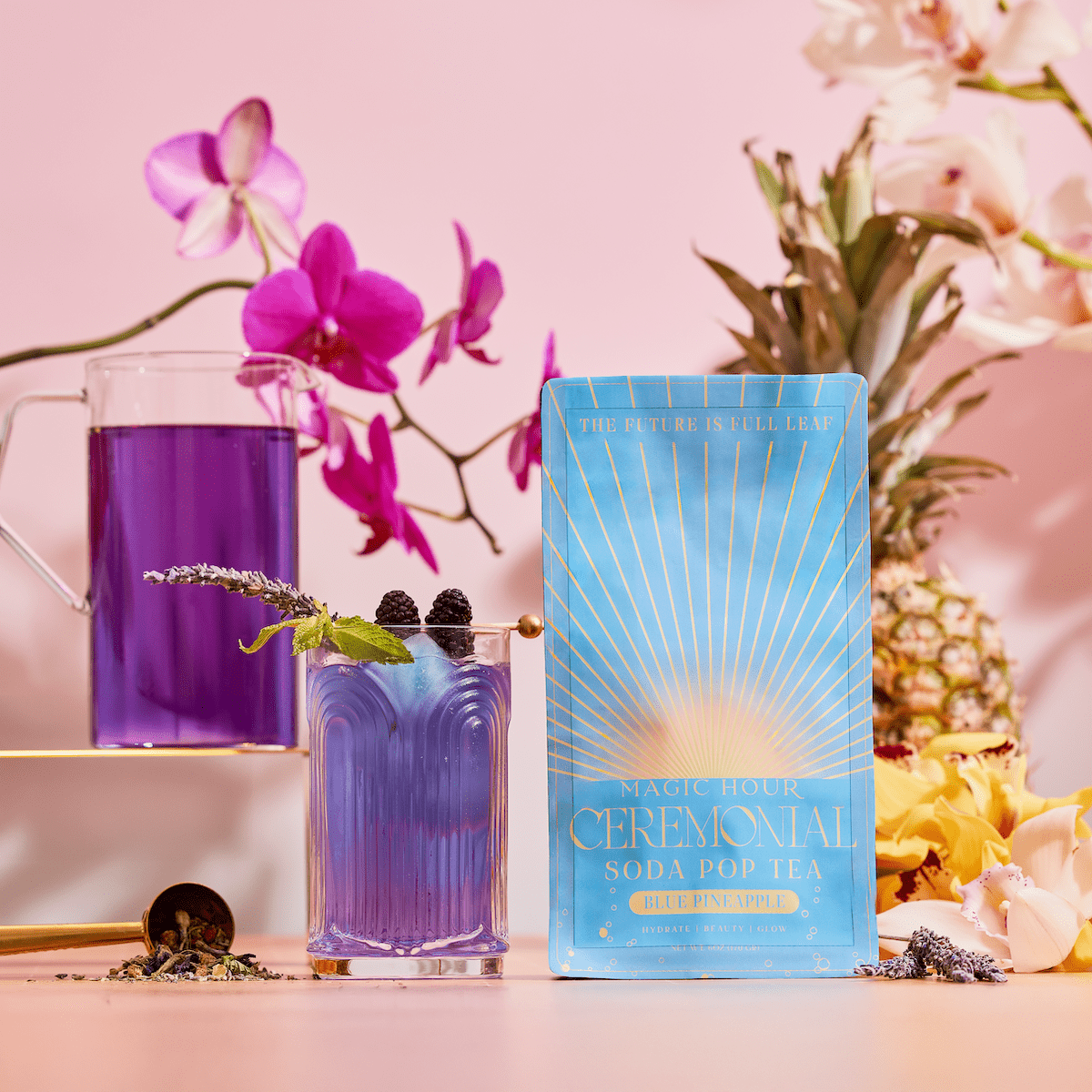A blue package of Blue Pineapple: Ceremonial Soda Pop Tea, crafted with antioxidant-rich tea and Blue Spirulina, sits before a glass and pitcher of vibrant purple tea with blackberries, lavender, orchids, pineapple, and yellow flowers on a pastel pink background.