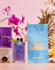 A blue package of Blue Pineapple: Ceremonial Soda Pop Tea, crafted with antioxidant-rich tea and Blue Spirulina, sits before a glass and pitcher of vibrant purple tea with blackberries, lavender, orchids, pineapple, and yellow flowers on a pastel pink background.