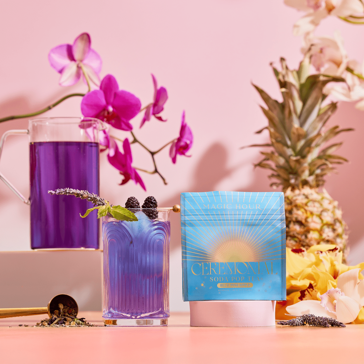 A blue cocktail with berries and a lavender garnish sits next to a Blue Pineapple: Ceremonial Soda Pop Tea box featuring Butterfly Pea Flower. The scene includes purple orchids, a pineapple, and yellow flowers on a pink background.