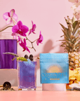 A blue cocktail with berries and a lavender garnish sits next to a Blue Pineapple: Ceremonial Soda Pop Tea box featuring Butterfly Pea Flower. The scene includes purple orchids, a pineapple, and yellow flowers on a pink background.