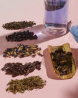 An assortment of dried herbs and organic blueberries arranged in small piles on a light pink surface. Next to them is a glass filled with Magic Hour Blueberry Cheesecake Oolong Iced Tea and a golden dish containing a herb blend. A blue object in a glass container is also visible.