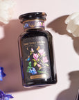 A dark-colored, elegantly designed tea canister labeled "Magic Hour - Blueberry Cheesecake Oolong Iced Tea" is surrounded by white tulips and blue flowers. The canister, featuring a whimsical illustration of flowers and organic blueberries, promises a delightful iced tea experience.