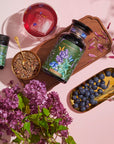 A vibrant arrangement featuring a large jar, a small bottle, a cup of Blueberry Lavender Mint: Cosmic Garden Iced Tea by Magic Hour with an ice cube, a bowl of loose leaf tea with a gold spoon, a bunch of purple flowers, and a gold tray of blueberries during the magic hour. The jars have botanical labels, and petals are scattered on a wooden board.
