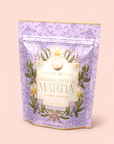 A lavender-colored pouch of Flower Powder: Blueberry Lavender Matcha with gold accents and floral designs, standing upright on a light pink background—perfect for those seeking a calming tea experience with the delicate flavors of blueberry matcha.