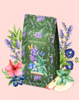 A Blueberry Lavender Mint: Cosmic Garden Iced Tea bag, adorned with lavender, butterfly, and star designs, is surrounded by illustrated lavender, hibiscus, pansy flowers, blueberries, mint leaves, and apple slices on a pale pink background—perfect for brewing an antioxidant-rich iced tea or savoring a refreshing cup of blueberry lavender mint tea.