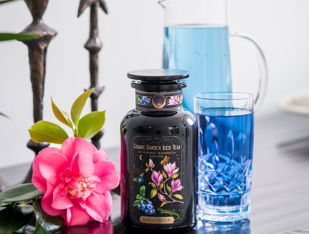 A black bottle labeled "Cosmic Garden Iced Teas Blueberry Magnolia" stands on a wooden table beside a blue drink in a glass. A glass pitcher containing the same blue liquid is in the background. A pink flower and green leaves are placed next to the bottle.