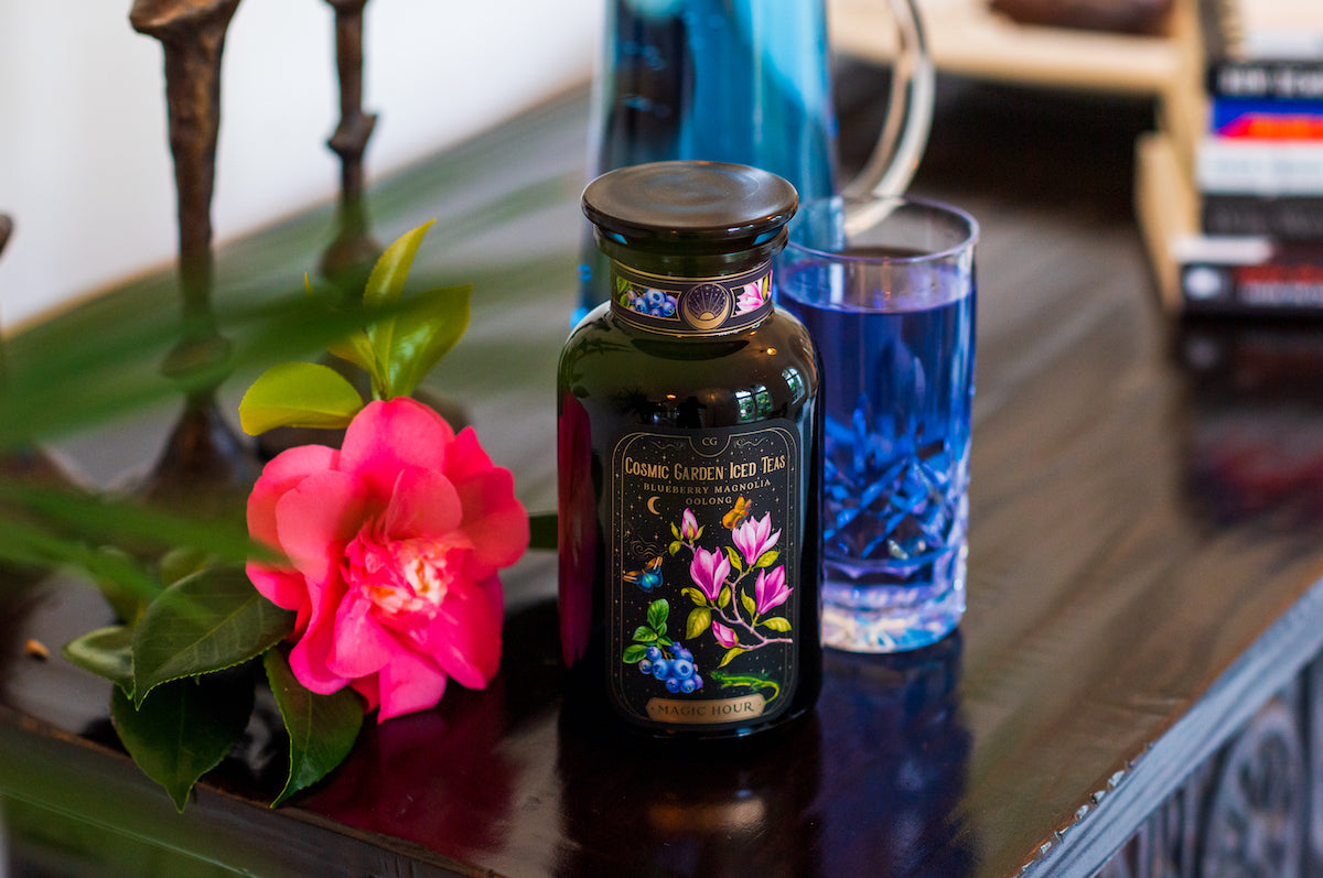 A black jar labeled "Cosmic Garden Iced Tea" is placed next to a blue flower and a vibrant pink flower on a dark wooden table. There's also a glass filled with blue liquid and a blue bottle in the background. Books are stacked to the right.