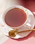 A Bohemian Breakfast Black Tea Bag steeps in a floral teacup on a matching saucer with a gold spoon, set atop a pink surface and soft white cloth—a stylish, organic coffee alternative.