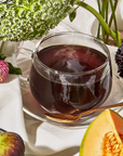 A glass cup of Bohemian Breakfast Black Tea sits on a white cloth with a saucer and gold spoon, surrounded by fresh flowers, fig, and cantaloupe. Steam rises from the tea, offering cozy comfort as a perfect coffee alternative.
