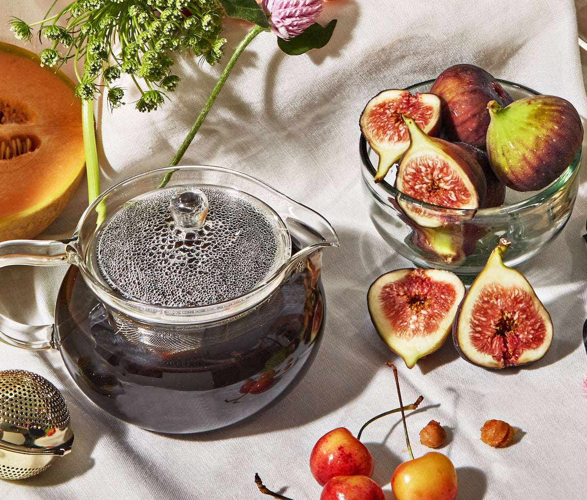 A clear glass teapot filled with dark tea sits on a white cloth next to a bowl of fresh figs, some halved. Nearby are a melon slice, cherries, and sprigs of flowers, creating a vibrant and fresh display.