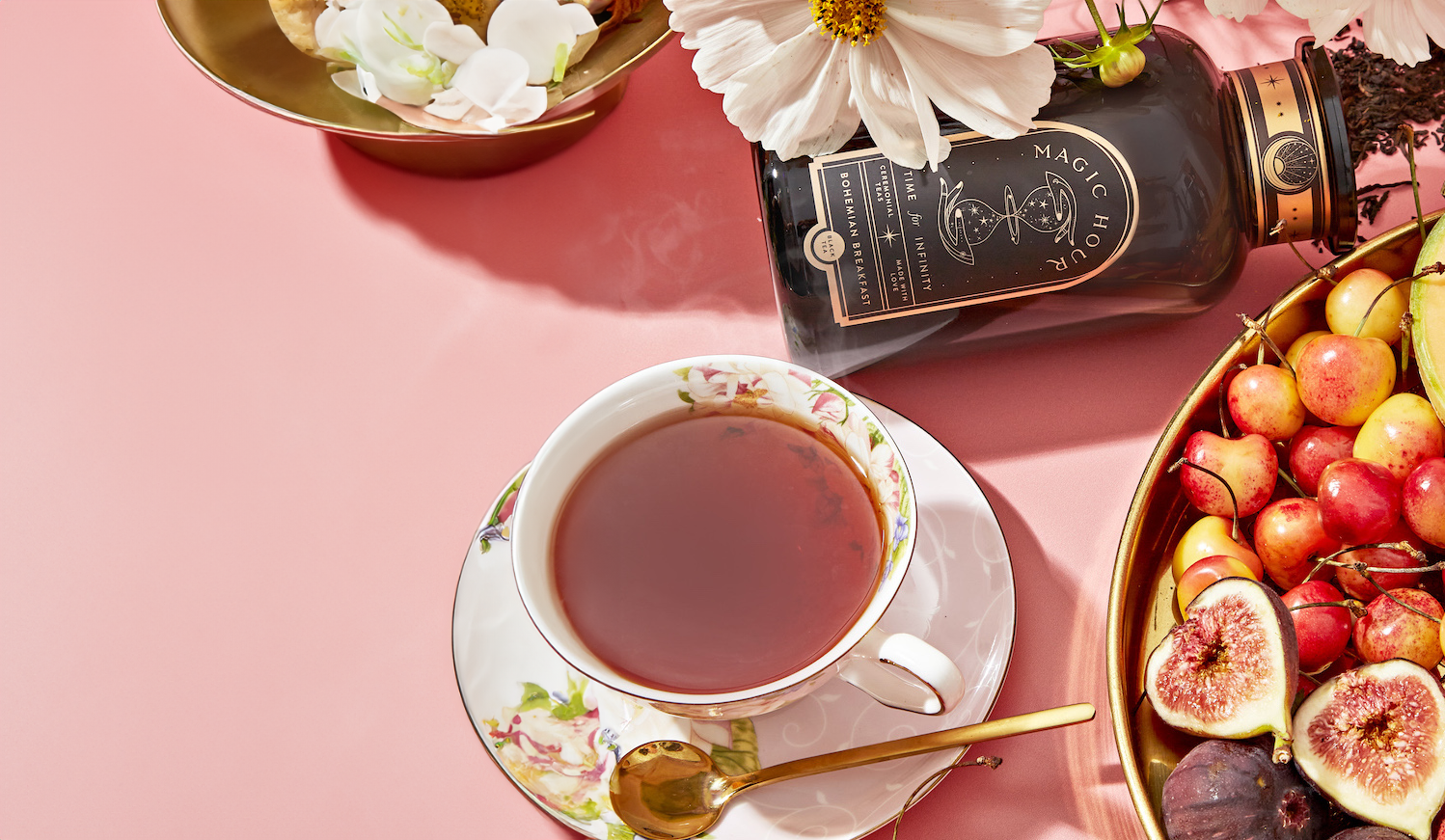 A cup of tea in a floral china teacup with a gold spoon rests on a pink surface. Nearby, there's a jar with a label containing text and a flower in the lid. Beside this is a bowl filled with assorted fruits including figs, cherries, and grapes.