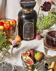 A dark apothecary jar of Best Sellers of Magic Hour Trio Box Bohemian Breakfast tea surrounded by a bowl of cherries, a bowl of sliced figs, a cup of tea, and pieces of cantaloupe.