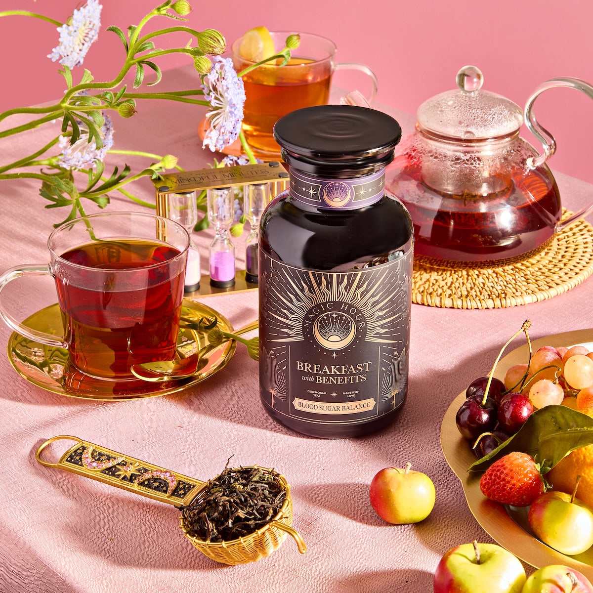 A jar of "Breakfast with Benefits: Blood Sugar Balance" is on a table amid teacups, a teapot, fresh grapes and strawberries, and a spoonful of Yunnan Black Tea. The bright scene features a pink background decorated with flowers.