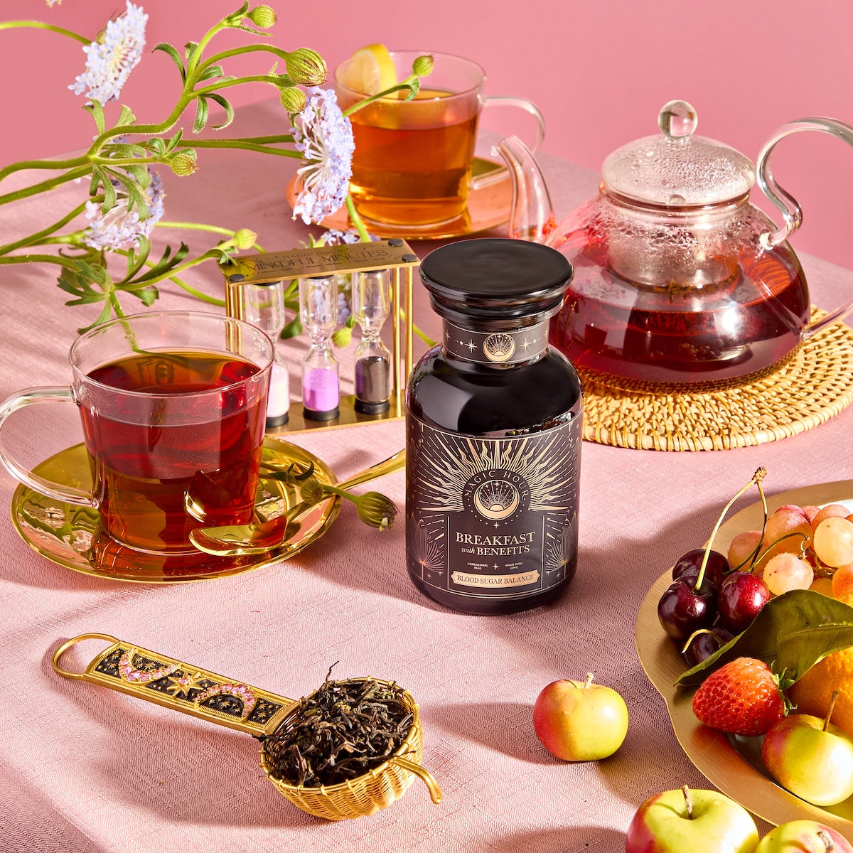 A breakfast scene features Yunnan Black Tea in gold-saucered cups, a glass teapot, and a "Breakfast with Benefits: Blood Sugar Balance" jar. Fresh flowers, a sand timer, cherries, apples on a pink tablecloth, and loose tea leaves in a scoop complete the setting.