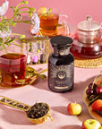 A breakfast scene features Yunnan Black Tea in gold-saucered cups, a glass teapot, and a "Breakfast with Benefits: Blood Sugar Balance" jar. Fresh flowers, a sand timer, cherries, apples on a pink tablecloth, and loose tea leaves in a scoop complete the setting.