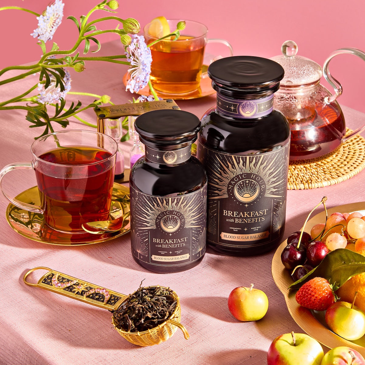 On a pink background, there's a set of ornate jars labeled "Breakfast with Benefits: Blood Sugar Balance," alongside a teapot and teacup, an Assam loose-leaf tea scoop, cherries, small apples, and flowers.