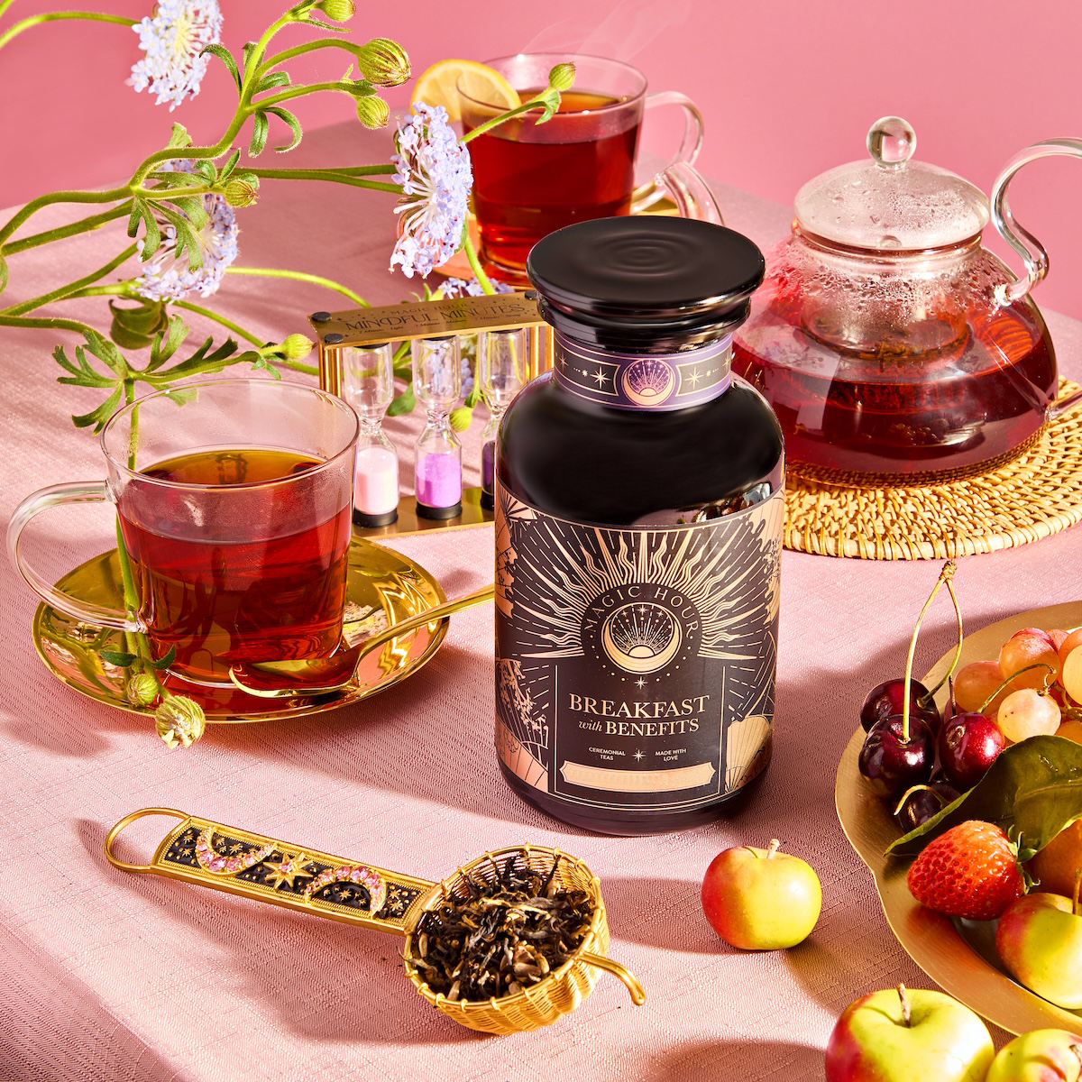 On a pink tablecloth, a jar of "Breakfast with Benefits: Metabolism" tea blend sits beside two glass teacups filled with black tea, a glass teapot, and a spoon of loose tea. Fresh flowers, cherries, and small apples enhance this vibrant, cozy scene.