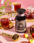 On a pink tablecloth, a jar of "Breakfast with Benefits: Metabolism" tea blend sits beside two glass teacups filled with black tea, a glass teapot, and a spoon of loose tea. Fresh flowers, cherries, and small apples enhance this vibrant, cozy scene.