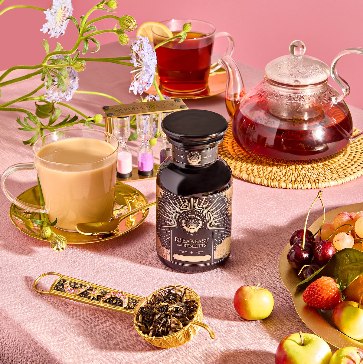 A set table showcasing the "Breakfast with Benefits: Metabolism" set includes fruit tea, a green tea-filled small teapot, a golden teacup of creamy tea, assorted mini fruits, a jar labeled "Breakfast Elements," a brass scoop with loose black tea, and decorative flowers on a pink backdrop.