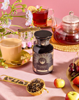 A set table showcasing the "Breakfast with Benefits: Metabolism" set includes fruit tea, a green tea-filled small teapot, a golden teacup of creamy tea, assorted mini fruits, a jar labeled "Breakfast Elements," a brass scoop with loose black tea, and decorative flowers on a pink backdrop.