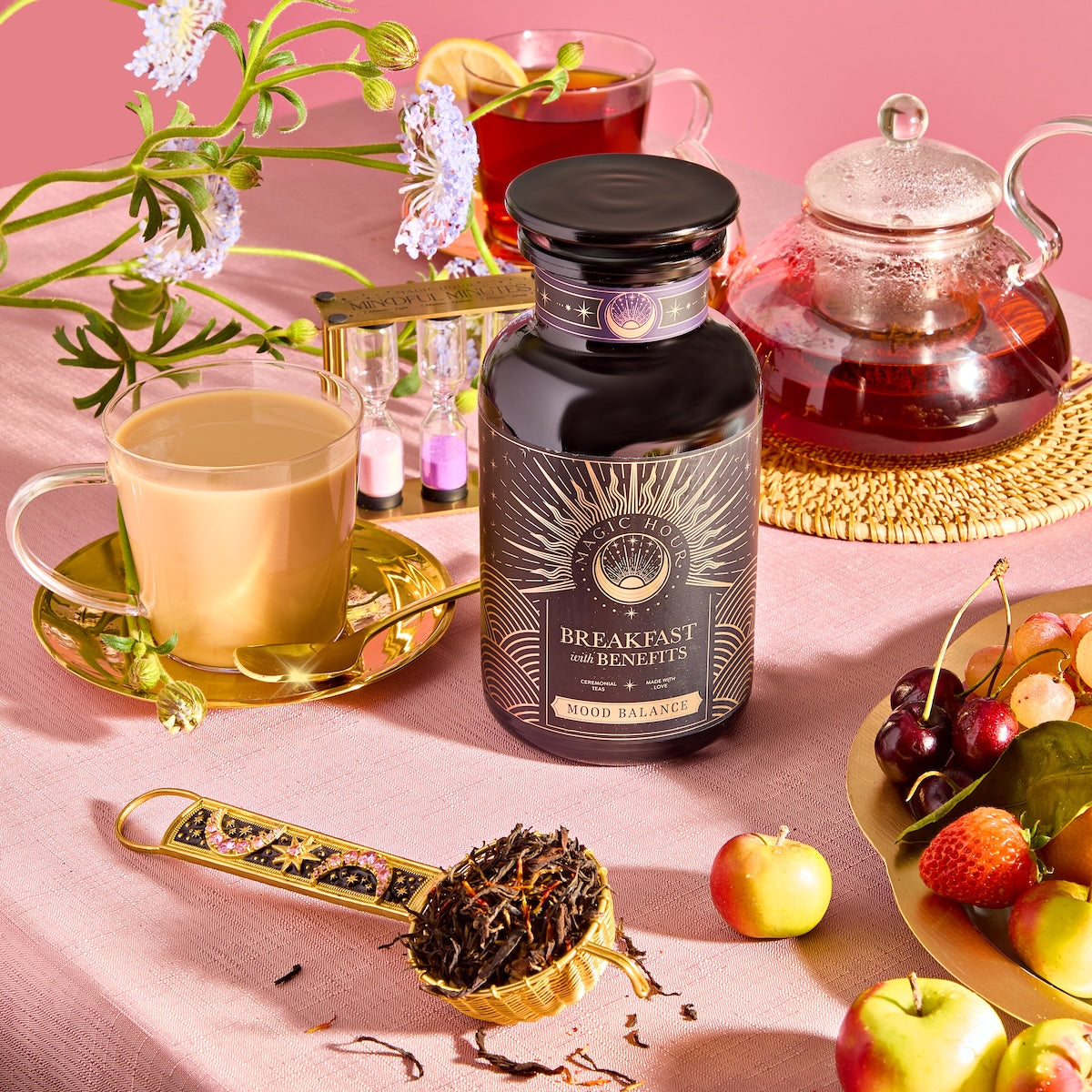 A "Breakfast with Benefits: Mood Lift" jar sits on a pink table with teacups, a teapot, and a spoon of loose Assam tea leaves. Flowers, an hourglass, and fresh fruit enhance the colorful and serene setting.