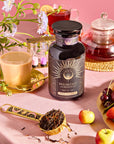 A "Breakfast with Benefits: Mood Lift" jar sits on a pink table with teacups, a teapot, and a spoon of loose Assam tea leaves. Flowers, an hourglass, and fresh fruit enhance the colorful and serene setting.