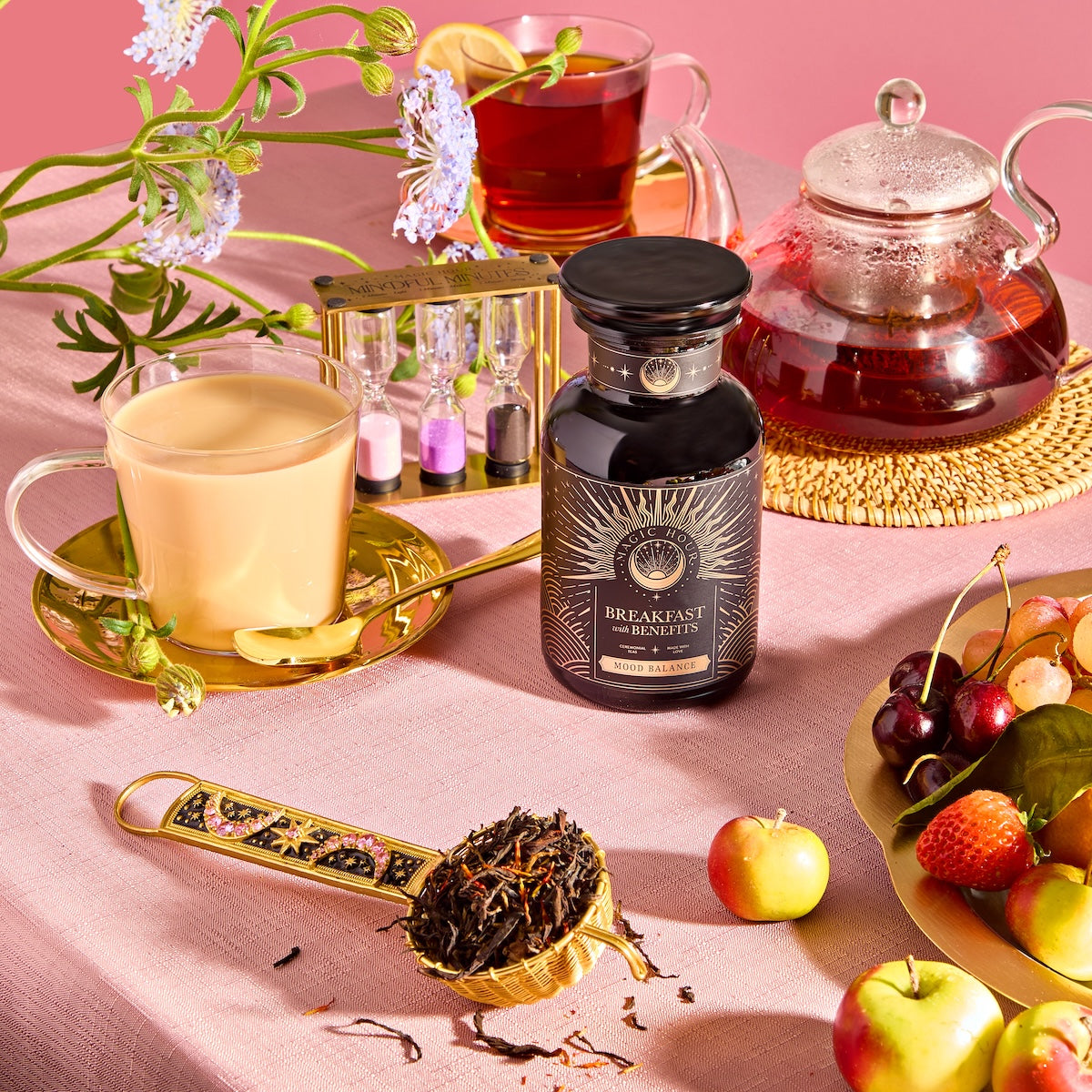 A lavish breakfast scene showcases a jar of "Breakfast with Benefits: Mood Lift" tea, a teapot, cups of milky and black Yunnan black tea, a sand timer, loose tea leaves on a spoon, and assorted fruits on the pink tablecloth. A flower adds an elegant touch to the arrangement.