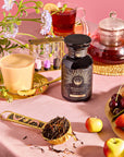 A lavish breakfast scene showcases a jar of "Breakfast with Benefits: Mood Lift" tea, a teapot, cups of milky and black Yunnan black tea, a sand timer, loose tea leaves on a spoon, and assorted fruits on the pink tablecloth. A flower adds an elegant touch to the arrangement.