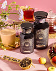 Two jars labeled "Breakfast with Benefits: Mood Lift" rest on a table beside Yunnan black tea, a teapot, a plate of fruit featuring cherries and grapes. Nearby is a small spoon with tea leaves, with flowers adorning the scene against a pink backdrop.