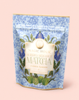 A blue floral-patterned pouch labeled “Flower Powder: Butterfly Bergamot Matcha”—featuring a blend of ceremonial matcha and butterfly pea flower—rests amid green leaves and limes on a pale pink background.