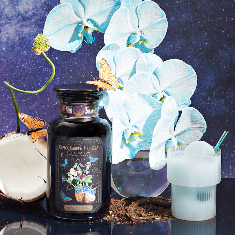 A dark bottle labeled "Cosmic Garden Iced Teas" is displayed with a drink in a frosted glass, white and blue orchids, a half-cut coconut, and butterflies. The background features a starry night sky, adding a mystical vibe to the arrangement.