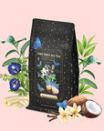 A packet of Butterfly Blue Coconut Cream Iced White Tea, adorned with a floral, butterfly, and starry design, is surrounded by fresh flowers, banana slices, vanilla, a cracked coconut, and green leaves on a soft pink background—perfect for crafting your own pina colada-inspired iced white tea or refreshing hydrating drink.