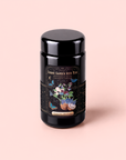A black tea canister labeled “Butterfly Blue Coconut Cream Iced White Tea.” The label features a floral arrangement with fruits, butterflies, stars, and decorative borders against a light pink background—perfect for storing your favorite hydration drink like blue tropical iced tea.