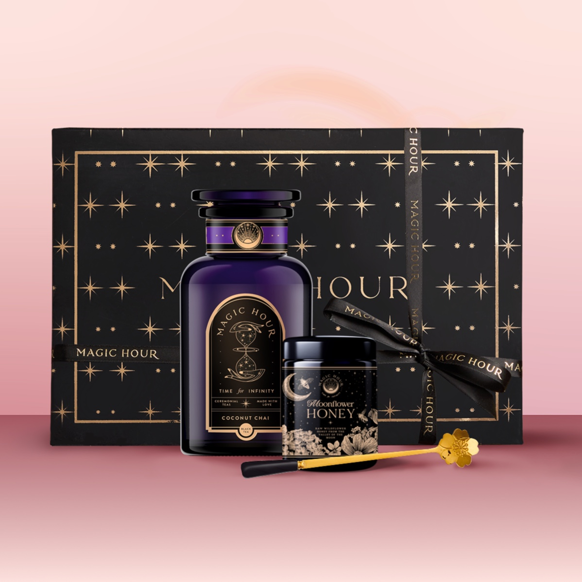 The Tea & Honey Gift Set is a luxurious morning gift, featuring a black "Magic Hour" box, purple coconut chai tea bottle, wildflower honey with golden dipper, and black ribbon, all arranged on a pink background.