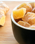 A bowl filled with sugar-coated pieces of Ginger Lemon sits on a wooden surface, with a piece of fresh ginger root in the background.
