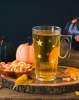 A glass mug with star designs holds yellow Candy Corn tea, set on a wooden slab beside a dish of Candy Corn, autumn leaves, and glittery black and orange pumpkins for a festive Halloween display.