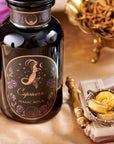 A black glass bottle labeled "Capricorn: Maple Oolong Tea" displays a mystical mermaid and celestial design, surrounded by gold objects and dried herbs, hinting at the tea's metabolism-boosting magic on an elegant surface.