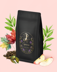 A bag of Capricorn: Maple Oolong Tea with a gold and purple Capricorn label stands on a pink background, surrounded by green leaves, apple slices, loose tea, metabolism-boosting cardamom pods, and autumn leaves.