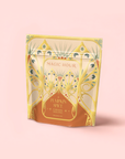 A decorative gold and orange stand-up pouch labeled "Pumpkin Spice Caramel Oolong" features an ornate, art nouveau-inspired design. This fall harvest pumpkin caramel tea is set against a soft pink background.